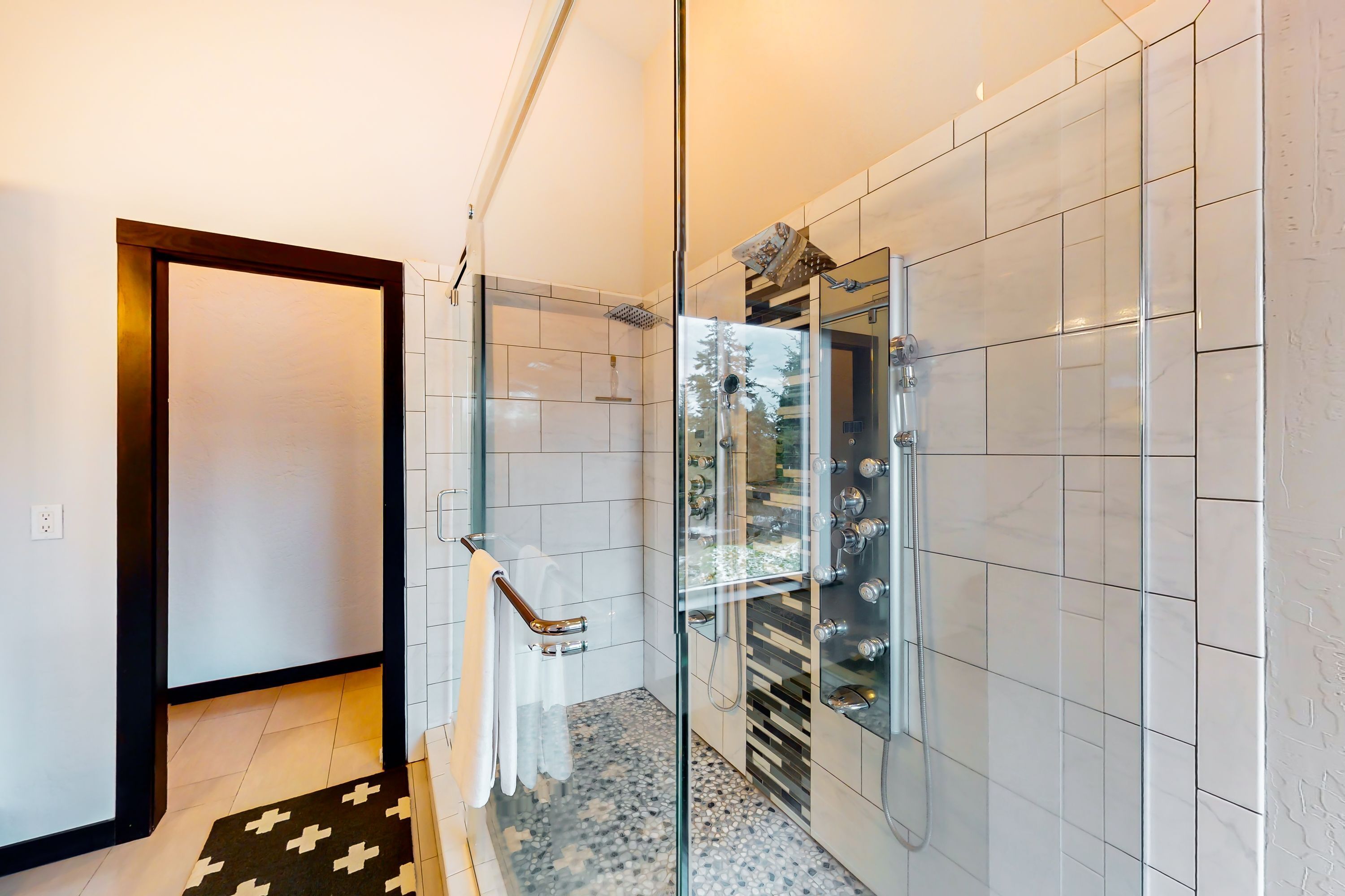 A modern bathroom with a glass shower enclosure, a towel rack, and a patterned floor mat.