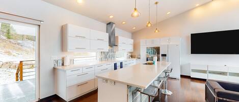 A modern kitchen with white cabinetry, a central island with bar stools, a large window, and a black TV mounted on the wall.