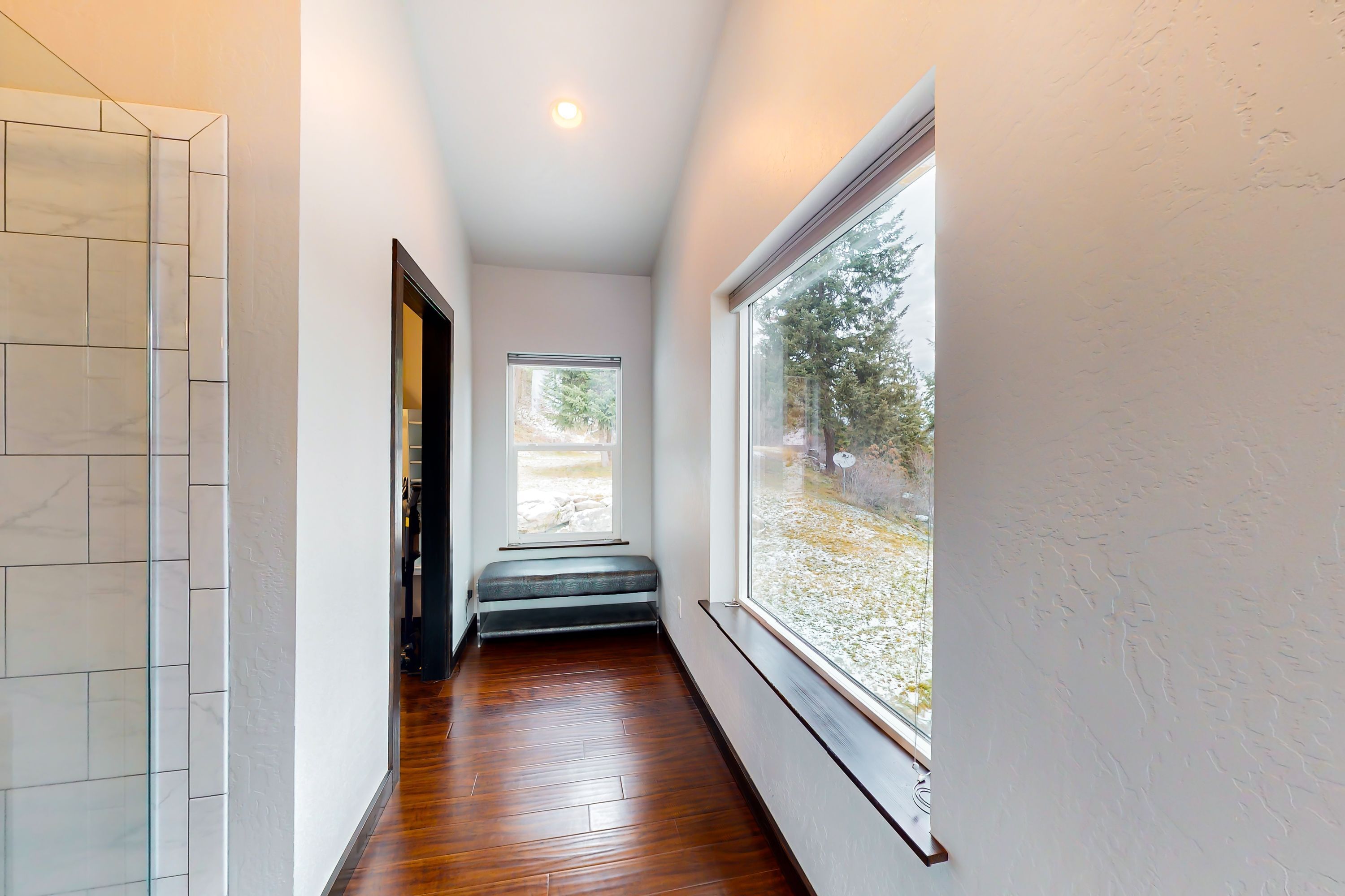 A narrow hallway with a wooden floor, a bench, a window with a view, and a tiled wall.