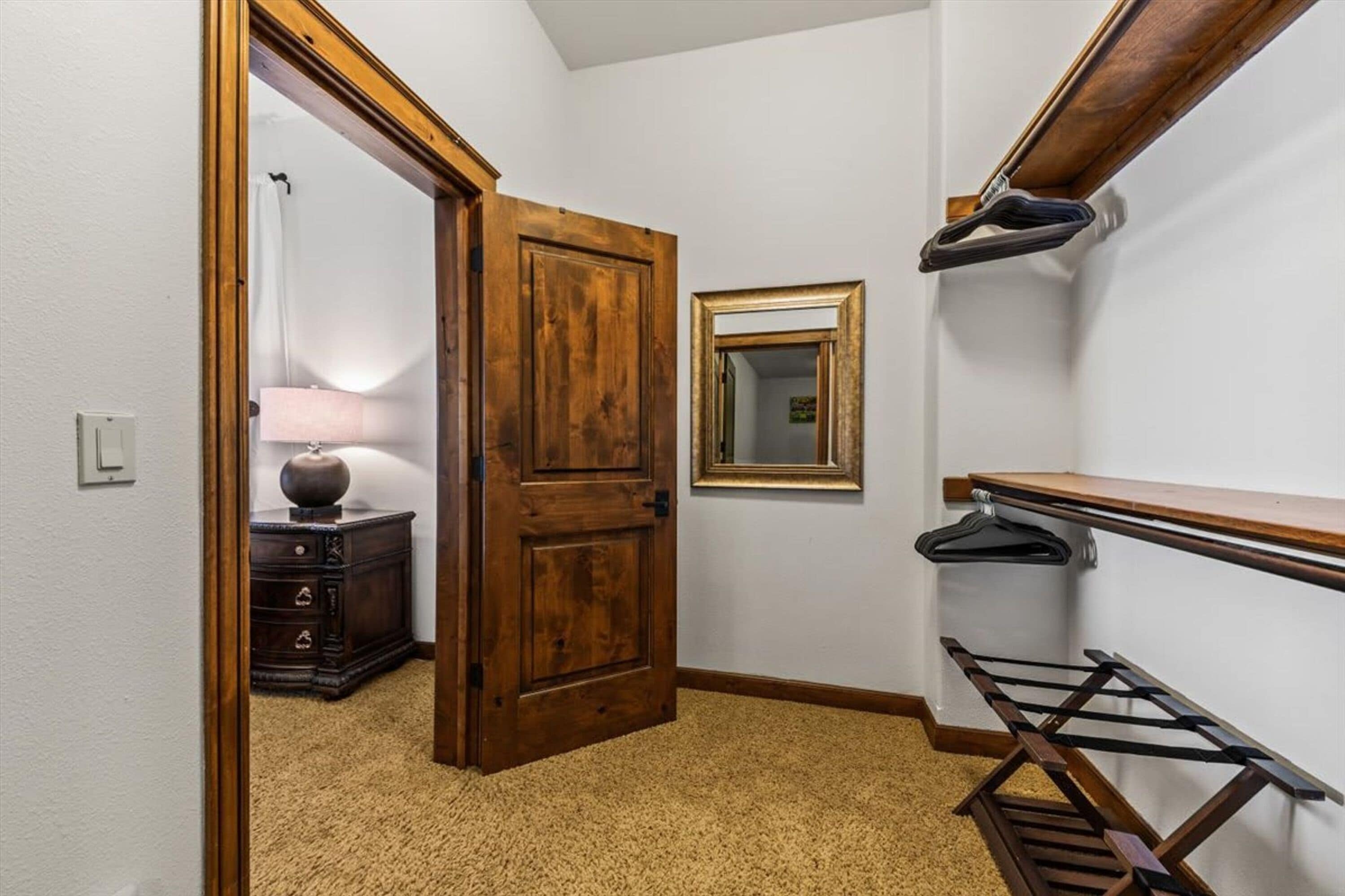 A hallway with a wooden door, a mirror, a lamp, and a wooden shelf with a hanging rack.