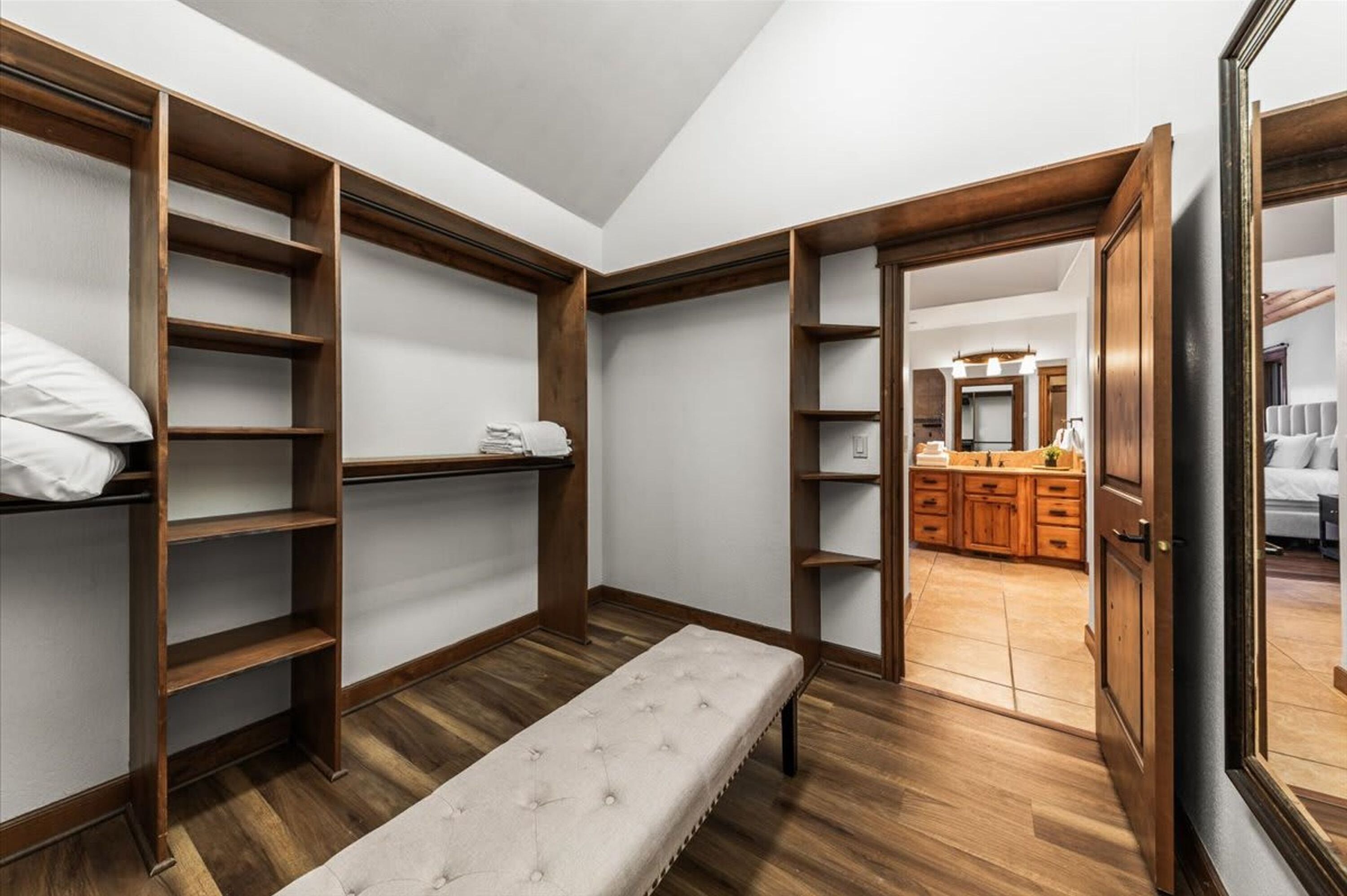 A bedroom with a wooden wardrobe, a bench, and a view into a kitchen area.