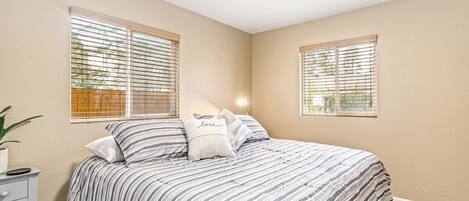 A bedroom with a bed, two windows, a bedside table, and a lamp.