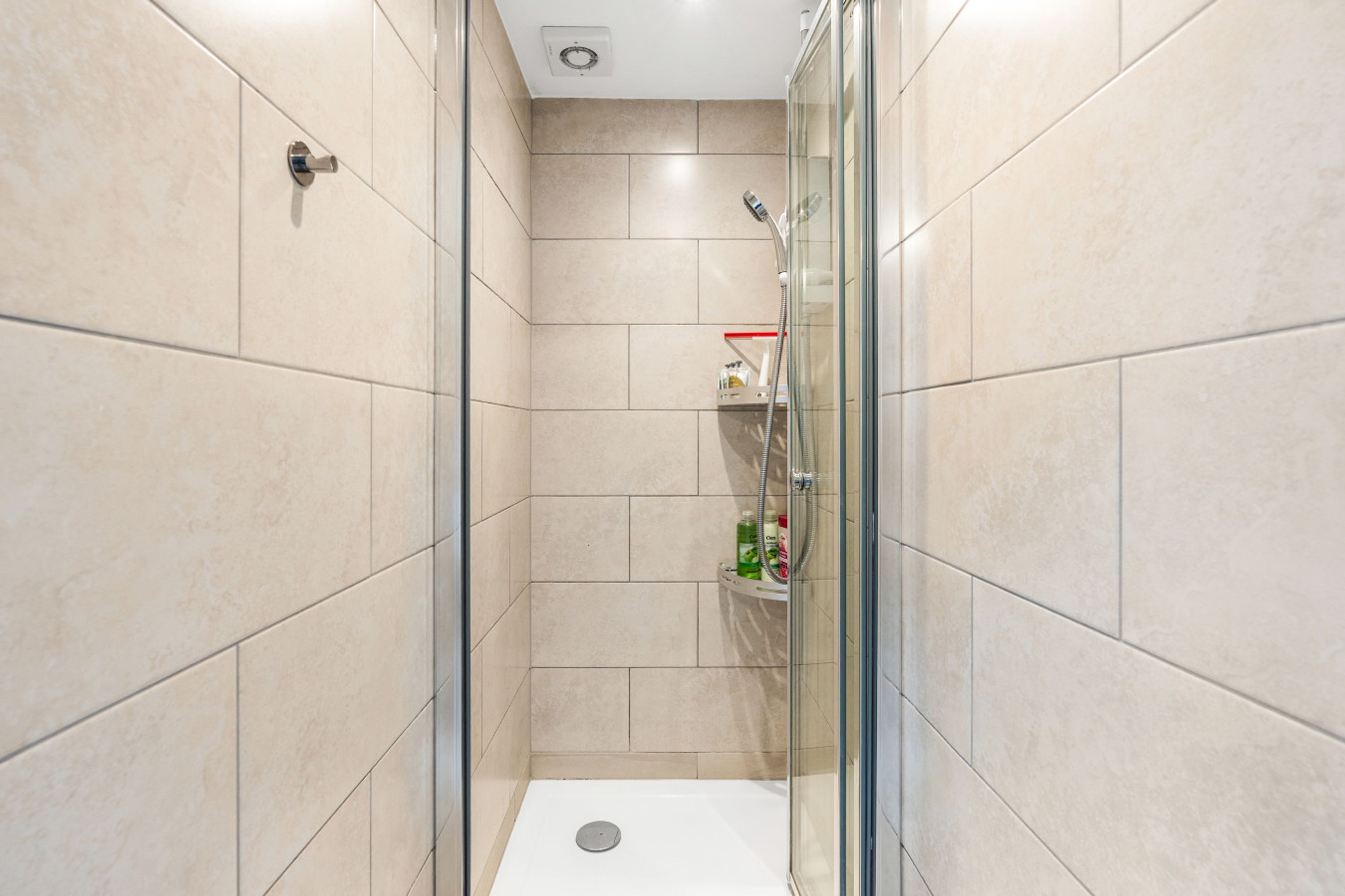 Une salle de bain moderne avec une douche à parois de verre, des carreaux de couleur beige et une petite étagère contenant des articles de toilette.