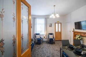 Une pièce comprenant une table à manger dressée pour quatre personnes, deux fauteuils, une petite table d’appoint, un téléviseur et une porte donnant sur une autre pièce.