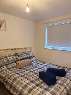 Un lit simple avec une couvre-lit à carreaux, une tête de lit et deux oreillers. Il y a une fenêtre avec des stores partiellement baissés, et une ampoule suspendue au plafond.