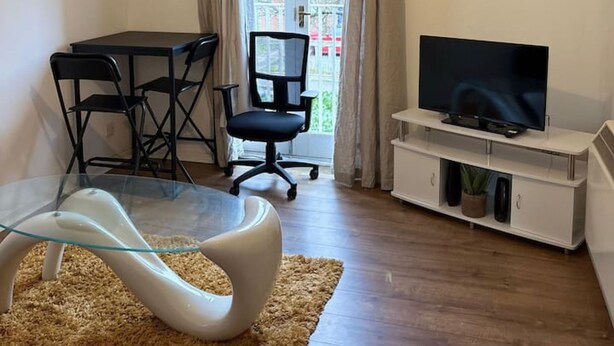 Een woonkamer met een glazen tafel, een zwarte bureaustoel, een televisie op een witte standaard en een raam met gordijnen.
