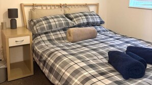 Een enkel bed met een geruit sprei, een houten hoofdbord, een nachtkastje met een lamp en een aan de muur hangend schilderij.