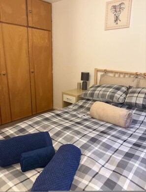 Une chambre avec une couvre-lit à carreaux, une armoire en bois, une table de chevet avec une lampe et un tableau accroché au mur.