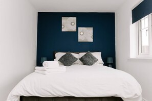 A neatly made bed with white linens, two pillows, and a stack of folded towels. There are two wall-mounted lamps on either side of the bed. - Hardwick Haven Modern 3BR Townhouse Retreat With Free Parking (Stockton-on-Tees)