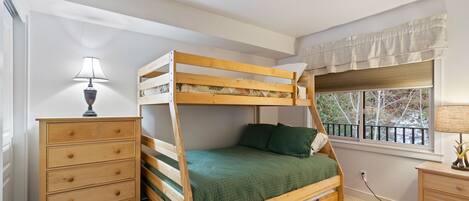 A bunk bed with a green bedspread, a wooden dresser, and a nightstand with a lamp.