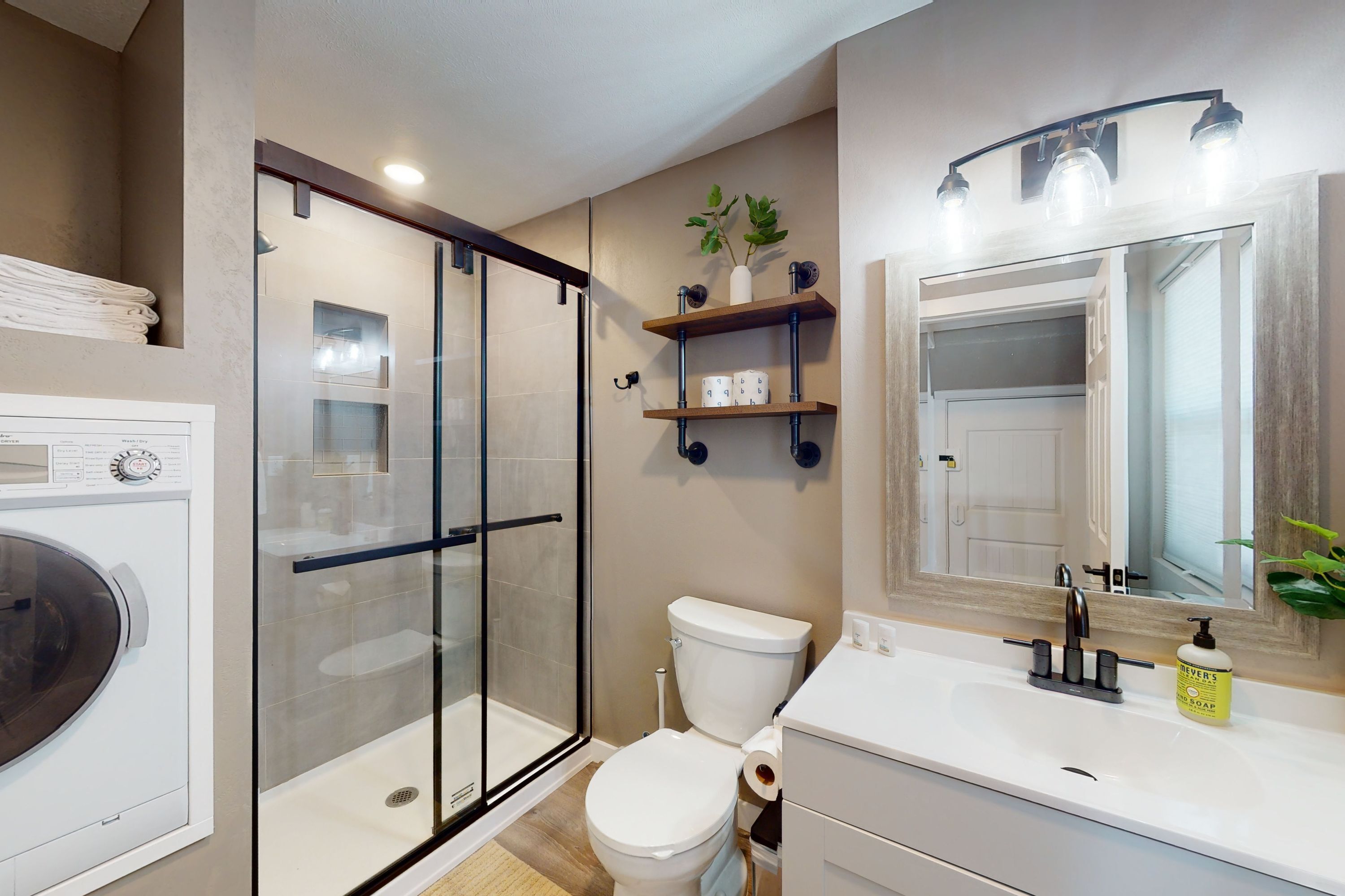 A bathroom with a glass shower enclosure, a toilet, a sink with a mirror, and a shelf with toiletries.