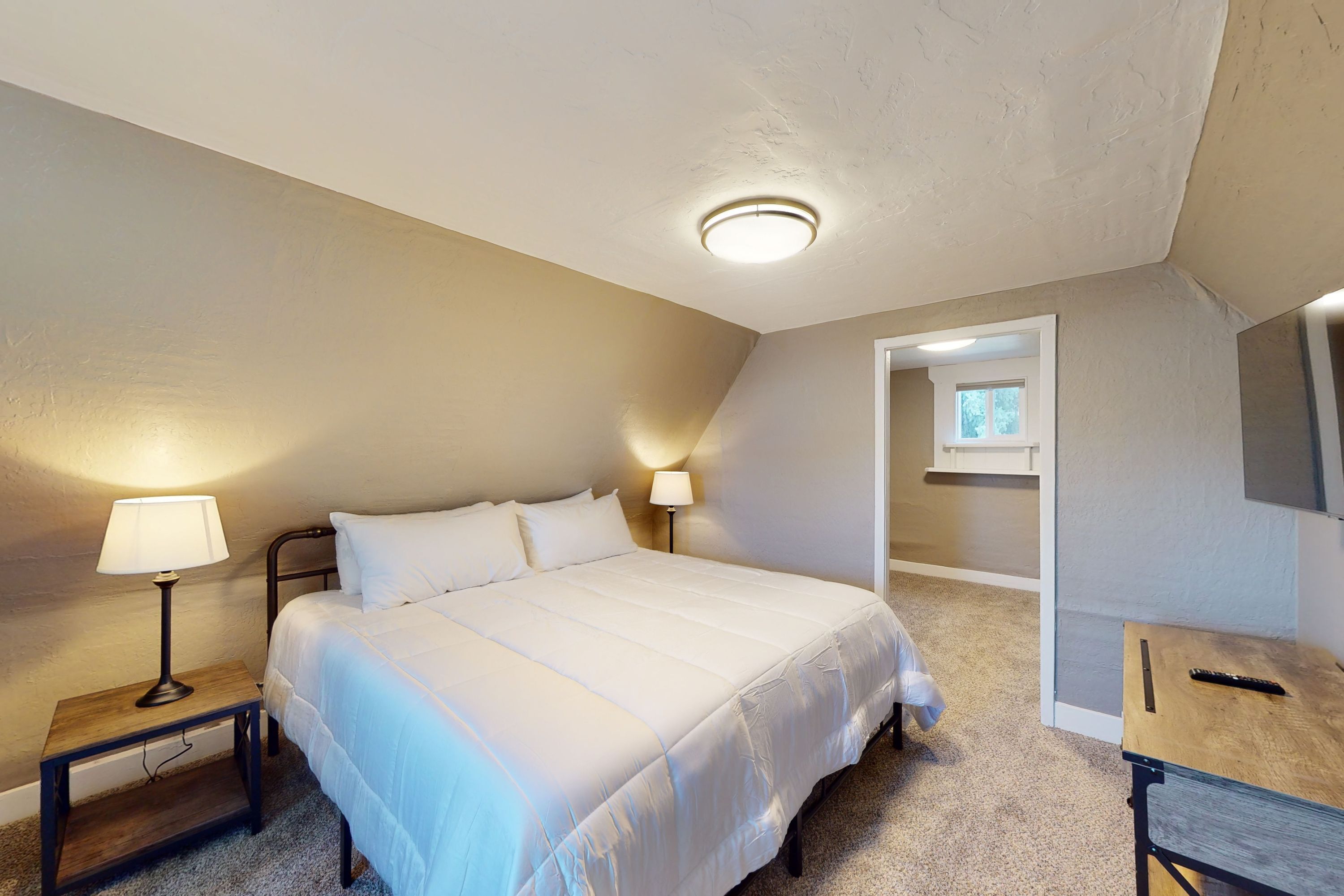 A bedroom with a bed, two bedside lamps, a wooden nightstand, a desk with a chair, and a ceiling light.