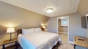 A bedroom with a bed, two bedside lamps, a wooden nightstand, a desk with a chair, and a ceiling light.