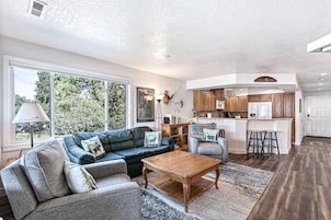 Amplia sala de estar con sofá, sillón y mesa de centro, una cocina con armarios de madera y un ventanal con vistas a árboles.