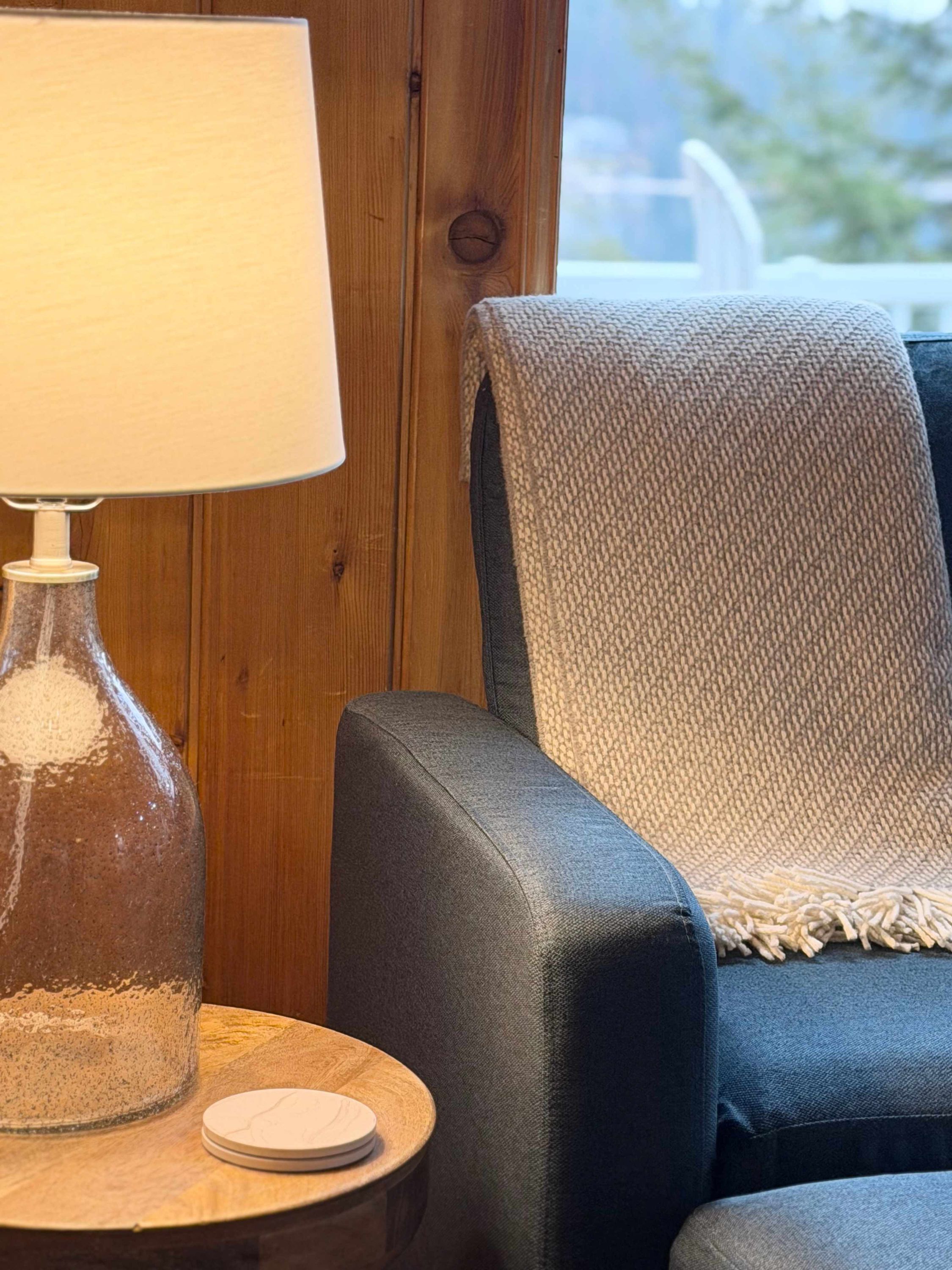 A room with a wooden wall, a lamp, a round table, and a blue chair with a grey blanket.