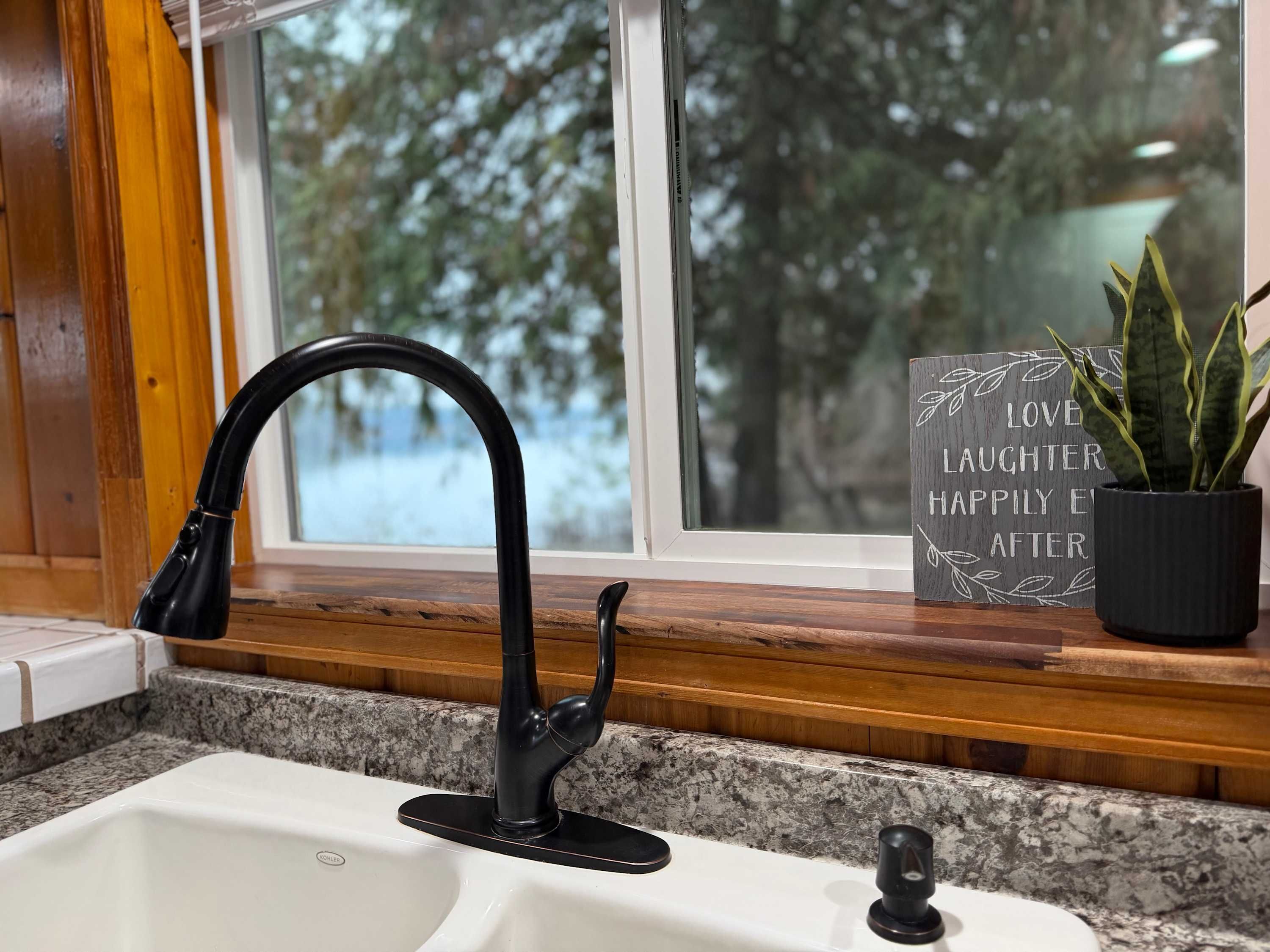 A kitchen with a black faucet, granite countertop, wooden window frame, and a view of trees outside.