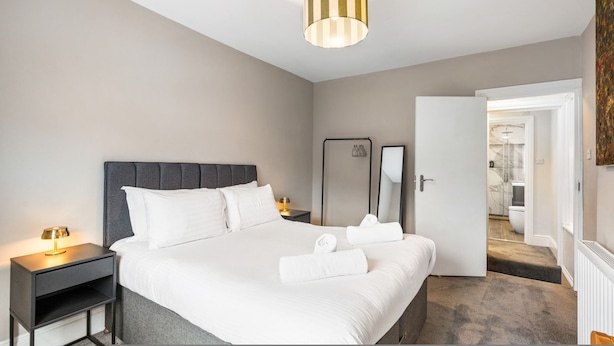 A hotel room with a bed, bedside tables, a mirror, and a door leading to a bathroom. - Spacious 5BR Home With Parking in Central Horsham (Horsham)
