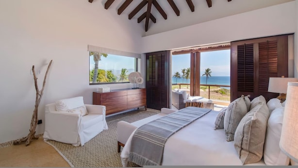 A bedroom with a bed, a white sofa, a wooden dresser, and a large window with a view of the ocean. - Casa Sole in Punta Mita (Punta de Mita)
