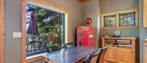 Un comedor con mesa y sillas, una ventana que da a vistas de vegetación, una máquina expendedora roja de Coca-Cola y armarios de madera.