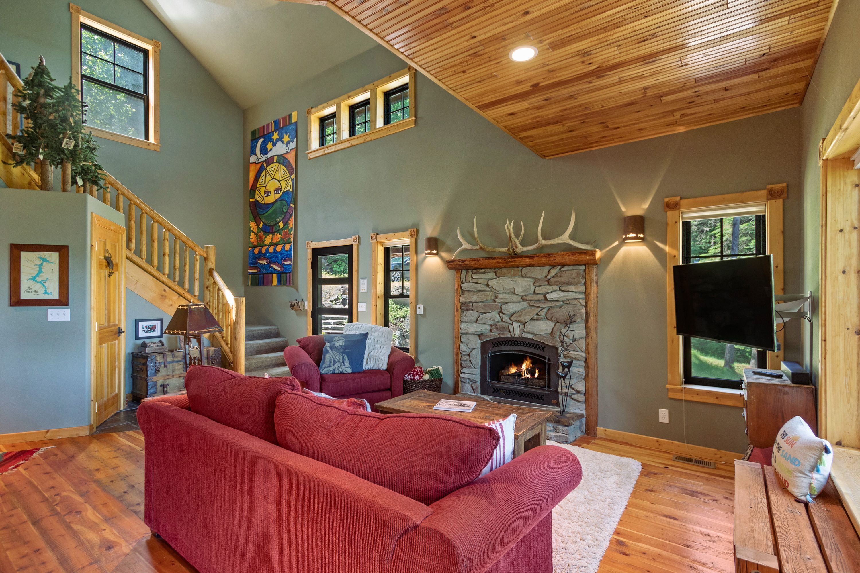 A cozy living room with a fireplace, a red sofa, a wooden staircase, and a stone fireplace surround.