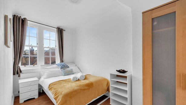Ein Schlafzimmer mit Bett, einem Fenster mit Vorhängen, einem Heizkörper, einem Kleiderschrank und einem Regal mit Gegenständen.