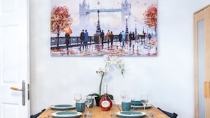 Ein Esstisch ist gedeckt mit Tellern, Gläsern und einer Flasche, an der Wand hängt ein Bild mit einer Brücke und Herbstlaub.