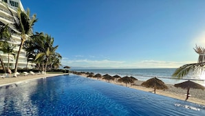 A clear blue pool with a view of the beach and ocean, surrounded by palm trees and lounge chairs. - Beautiful Beachfront 3 bd Condo in Nuevo Vallarta (Nuevo Nayarit)