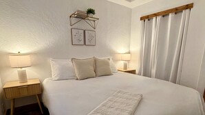 Un lit bien fait, recouvert d’une couette blanche, avec deux lampes de chevet, une plante sur la table de chevet et une étagère murale ornée de livres et d’une petite plante.