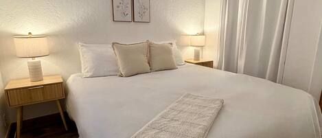 A neatly made bed with a white comforter, two bedside lamps, a bedside plant, and a wall-mounted shelf with books and a small plant.