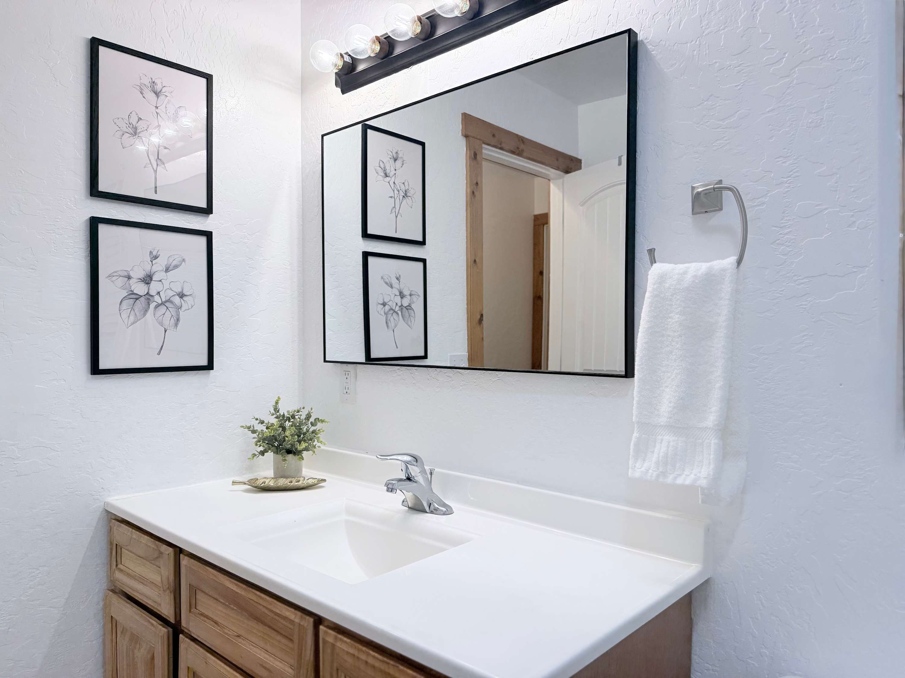 A bathroom with a white sink, wooden cabinets, a mirror with a light fixture, and framed floral artwork on the wall.