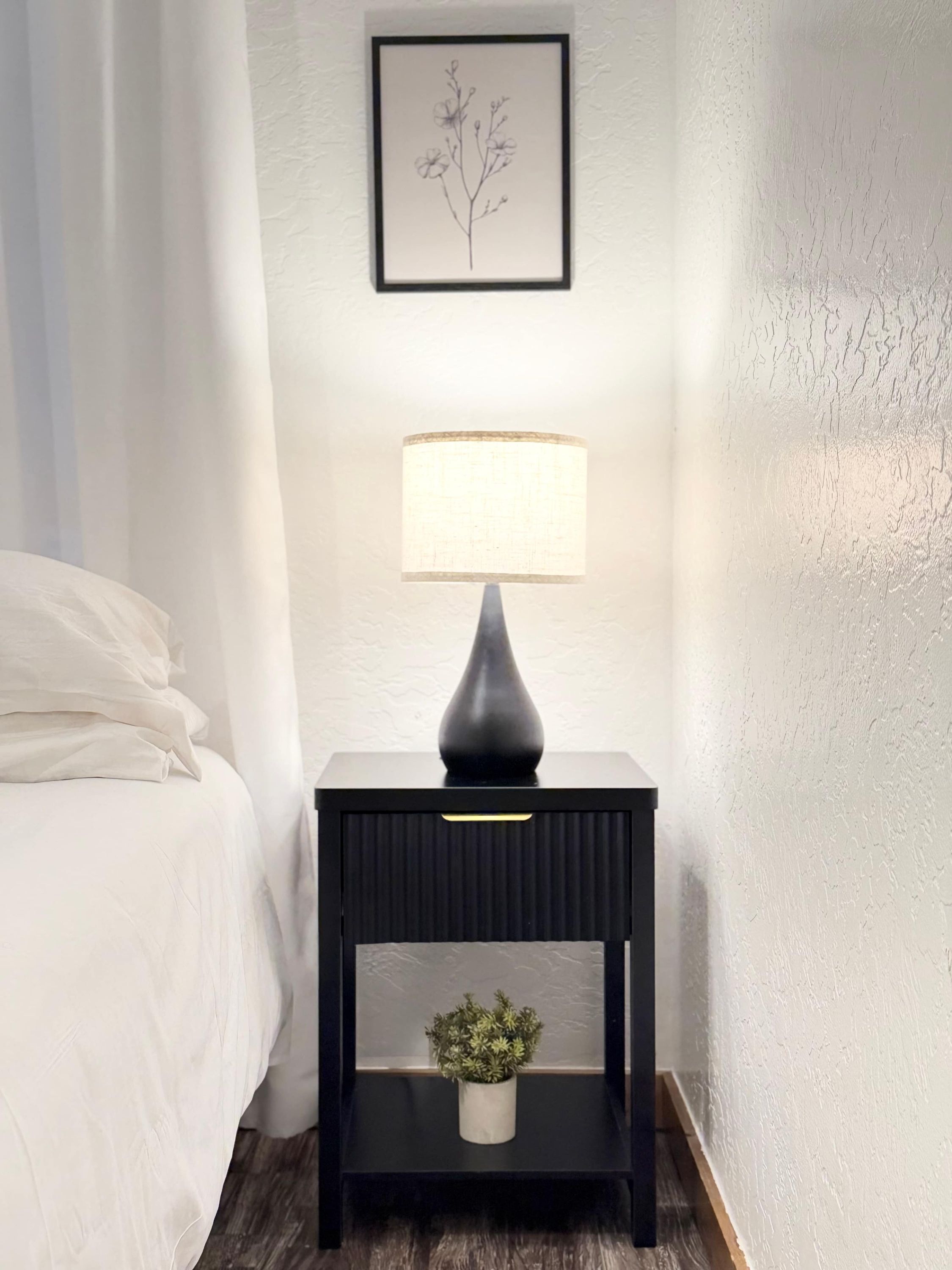A neatly made bed with white linens, a black bedside table with a lamp, a small shelf with a plant, and a framed botanical illustration on the wall.