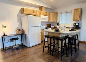 A modern kitchen with wooden cabinets, a white refrigerator, a microwave, a coffee maker, and a small island with stools. - Coeur d Alene Charm in Coeur d Alene (Coeur d'Alene)