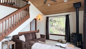 A cozy living room with a wooden staircase, a sofa set, a fireplace, and a large window with a view of greenery.