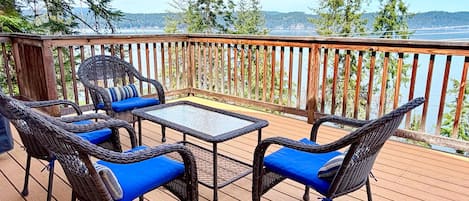 Eine Holzterrasse mit Korbmöbeln und einem Glastisch, mit Blick auf ein Gewässer und bewaldete Hügel.