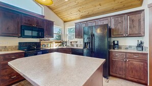 A kitchen with dark wooden cabinets, a large island, a black refrigerator, and a yellow pendant light.