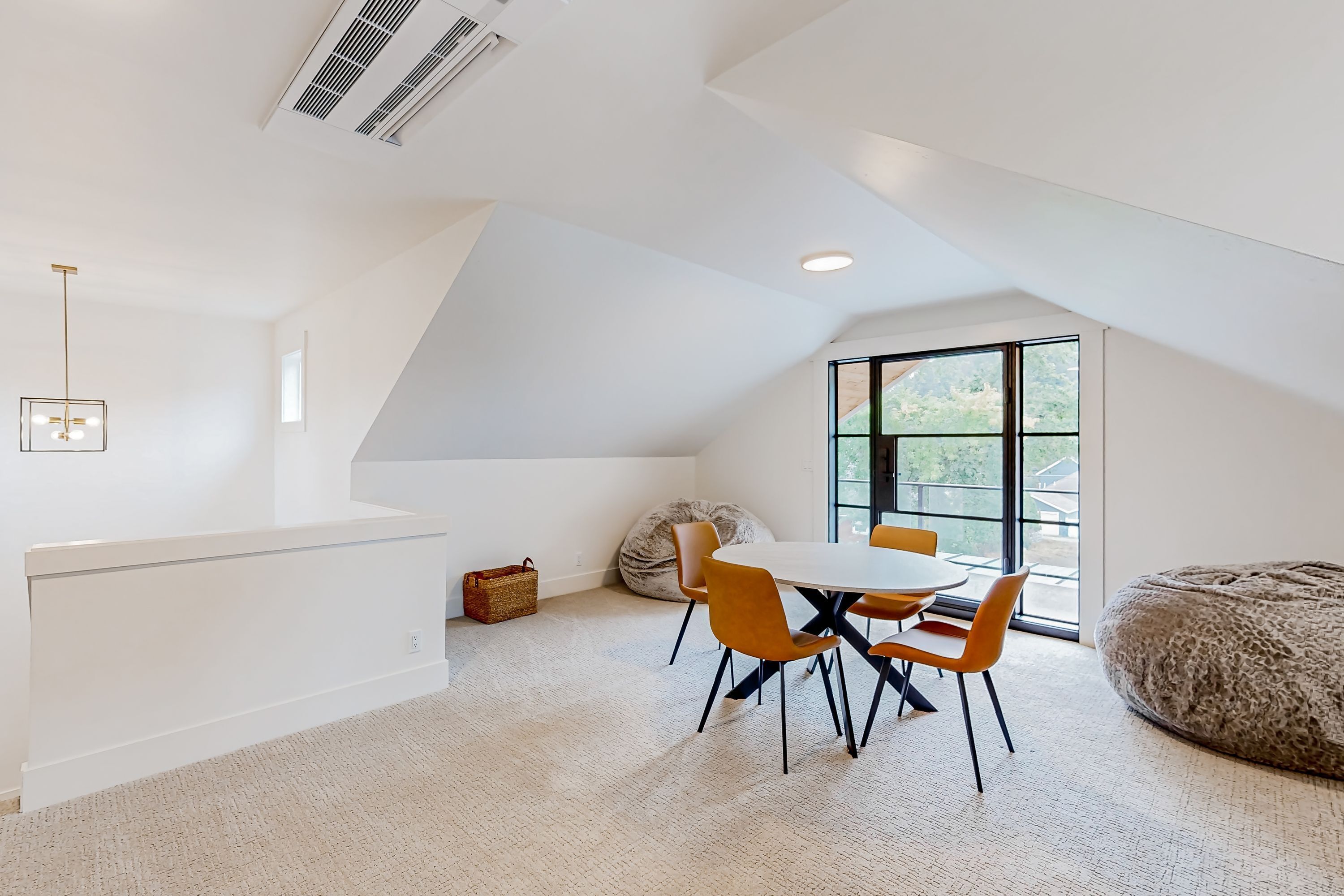 A modern loft bedroom with a sloped ceiling, a large window, a dining area with a round table and chairs, and a bean bag chair.
