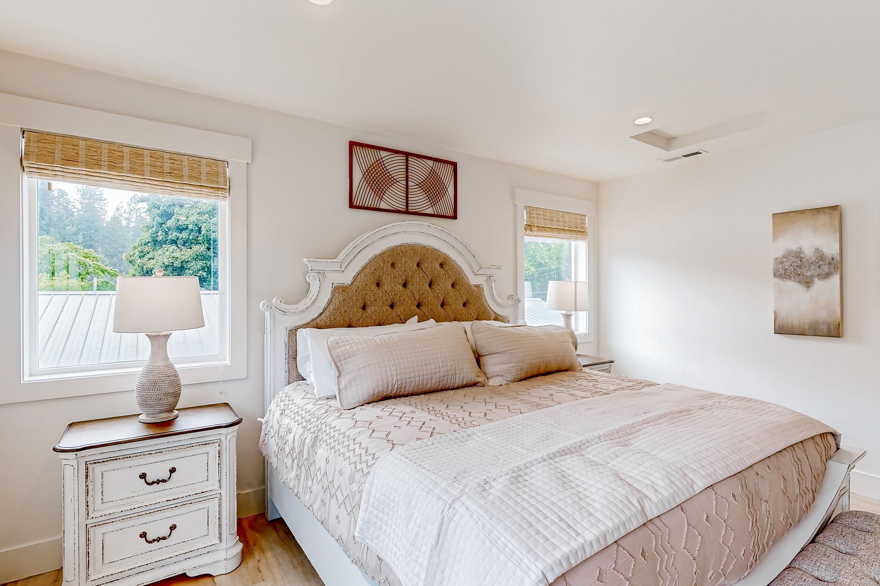 A bedroom with a large bed, a nightstand with a lamp, and a window with a view of greenery.