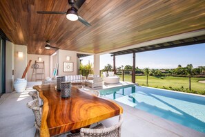 A modern outdoor dining area with a wooden table, wicker chairs, and a view of a swimming pool and golf course. - 3bed Condo- Private Pool Golf Cart Ocean View HK Las Marietas F101 (Punta de Mita)