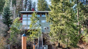 A two-story cabin with a dock extending into a river, surrounded by dense forest.
