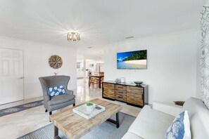 Ein Wohnzimmer mit einer Couch, einem Holztisch, einem gemusterten Sessel, einer Essecke mit Tisch und Stühlen, einem an der Wand befestigten Fernseher und einer Wanddekoration.