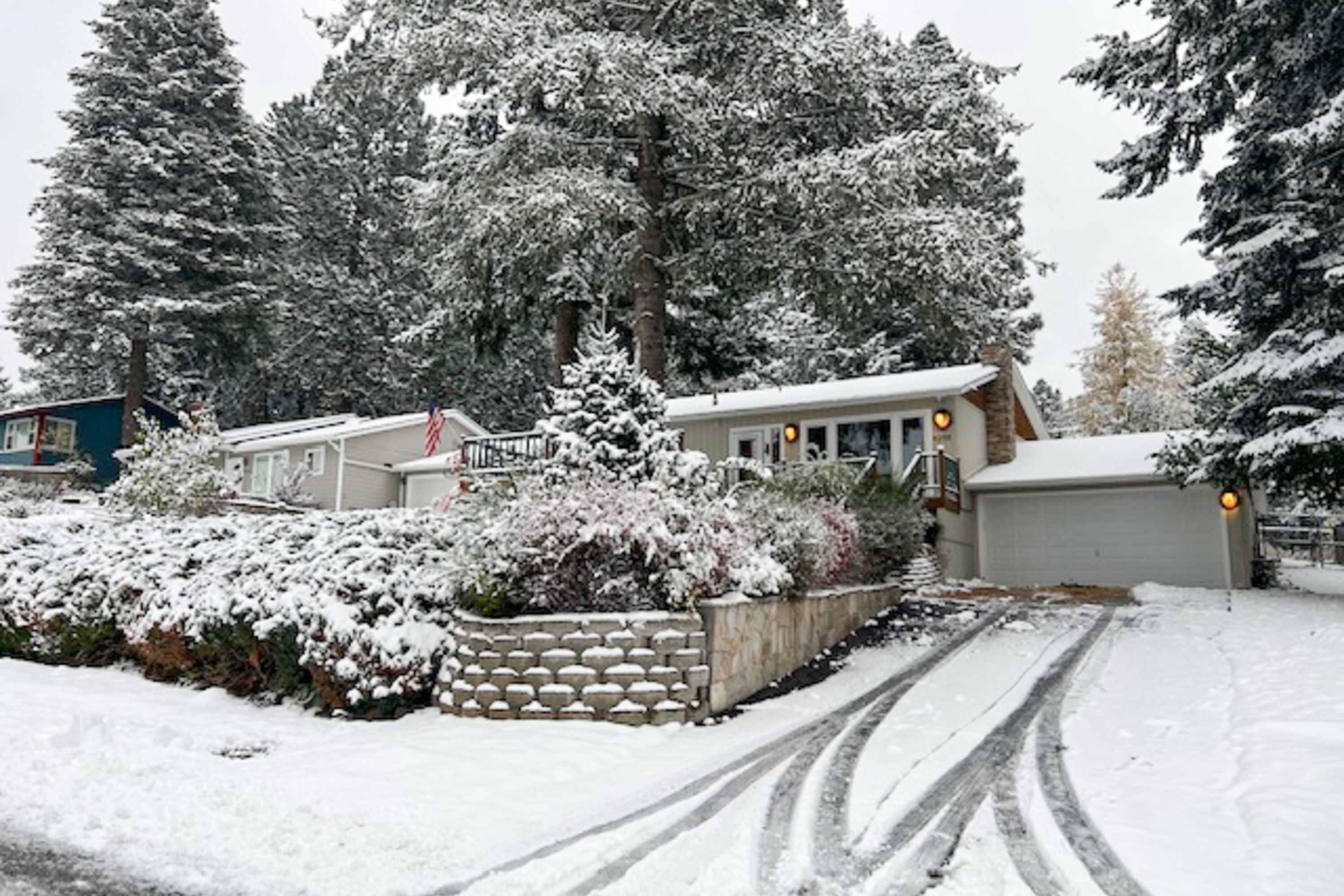 A snow-covered residential area with a house, a driveway, and snow-covered trees.