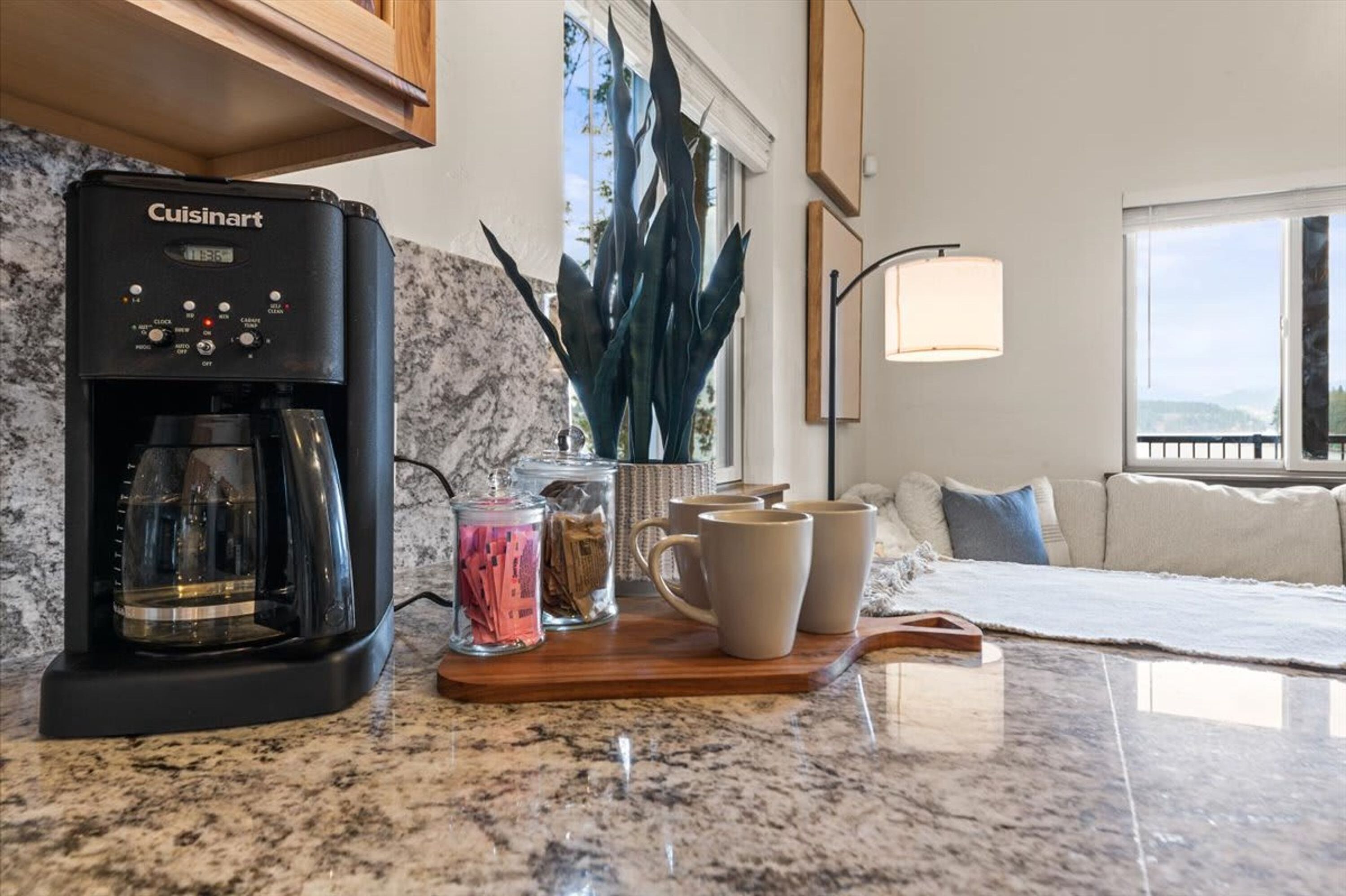 A kitchen with a Cuisinart coffee maker, a marble countertop, a wooden serving tray with cups and jars, and a view of a balcony through a window.