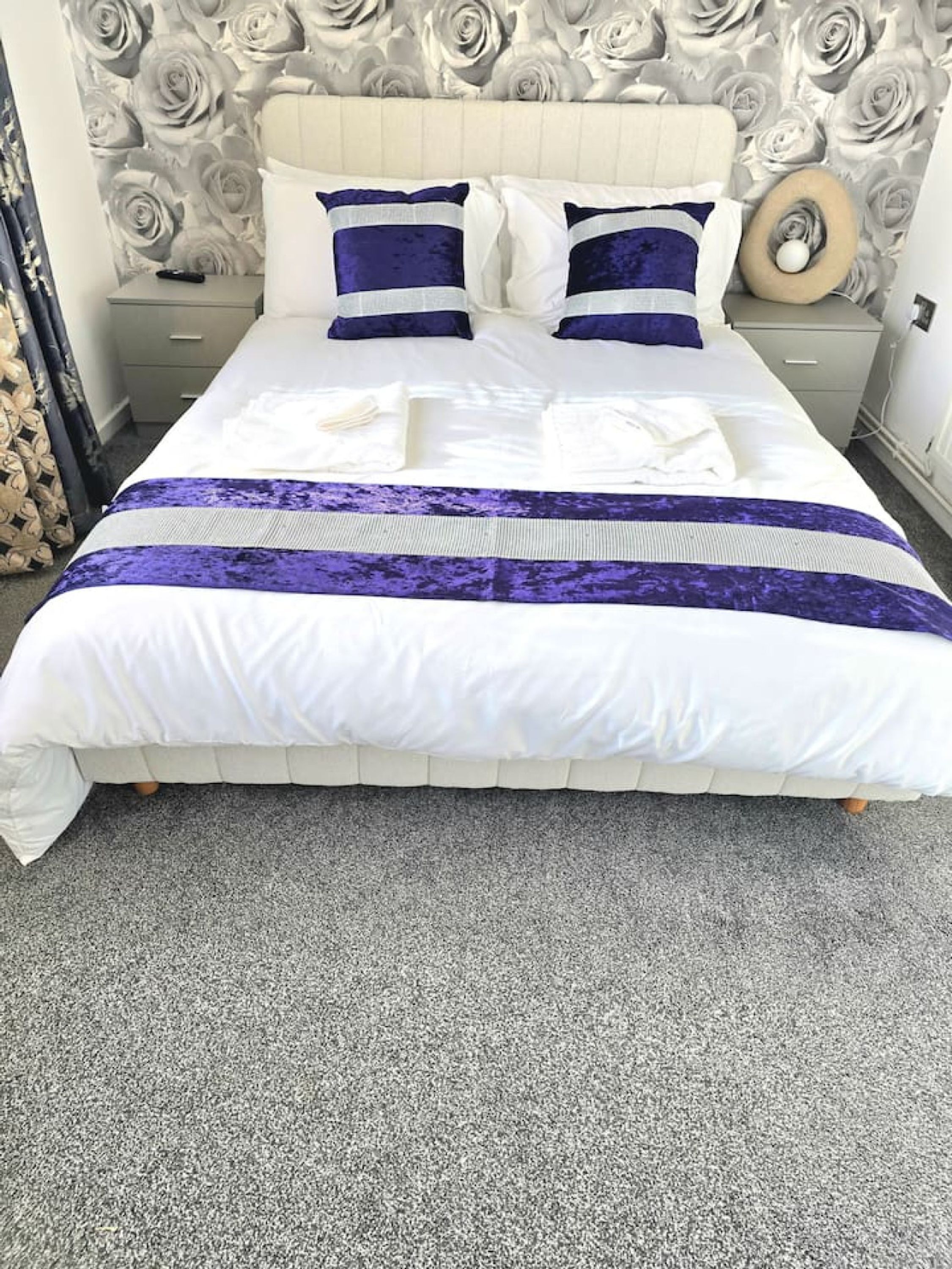 Ein ordentlich bezogenes Bett mit einer weißen und violetten Tagesdecke, ein Kopfteil mit einem Blumenmuster und ein grauer Teppich.
