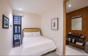 A bedroom with a bed, a nightstand, a window with blinds, a framed picture on the wall, and a bathroom with a sink and mirror. - Naya 1202 Tower I in Punta de Mita (Punta de Mita)