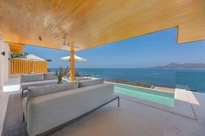 A modern living area with a sofa, a small table, and a plant. There is a pool with clear water and a view of the ocean and mountains in the background. - 4 bd Beach Chic Villa Ocean View - Iyari B4 (Punta de Mita)