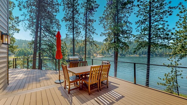 Une terrasse en bois avec une table et des chaises, donnant sur un lac bordé d’arbres et offrant une vue lointaine sur une maison.