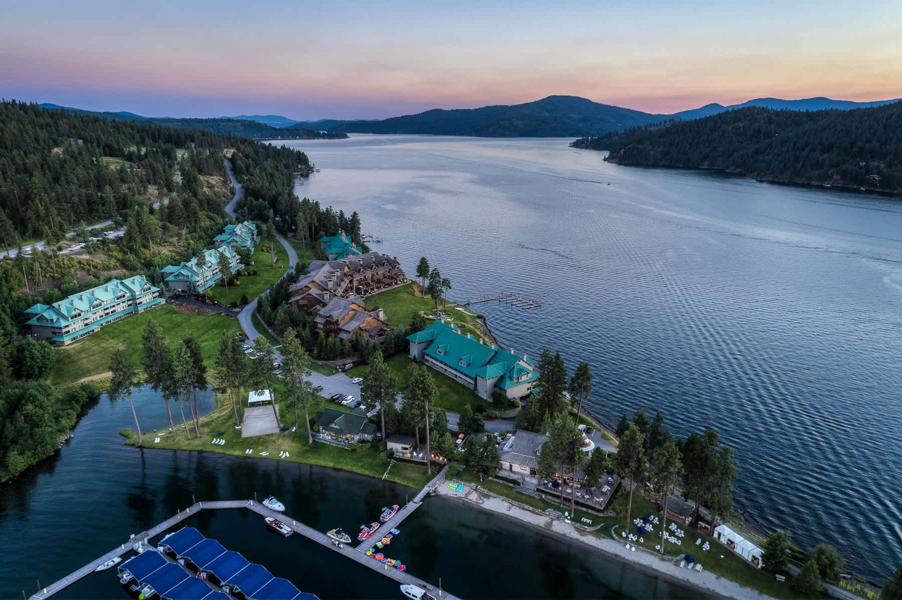 An aerial view of a lakeside resort with multiple buildings, a dock with boats, and a clear view of the lake.