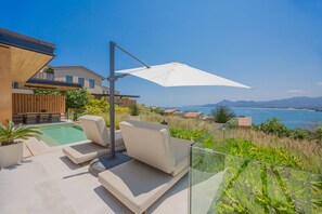 A poolside area with lounge chairs and a white umbrella, overlooking a body of water and hills. - 4 Bed - Luxury Oceanview Villa (Punta de Mita)