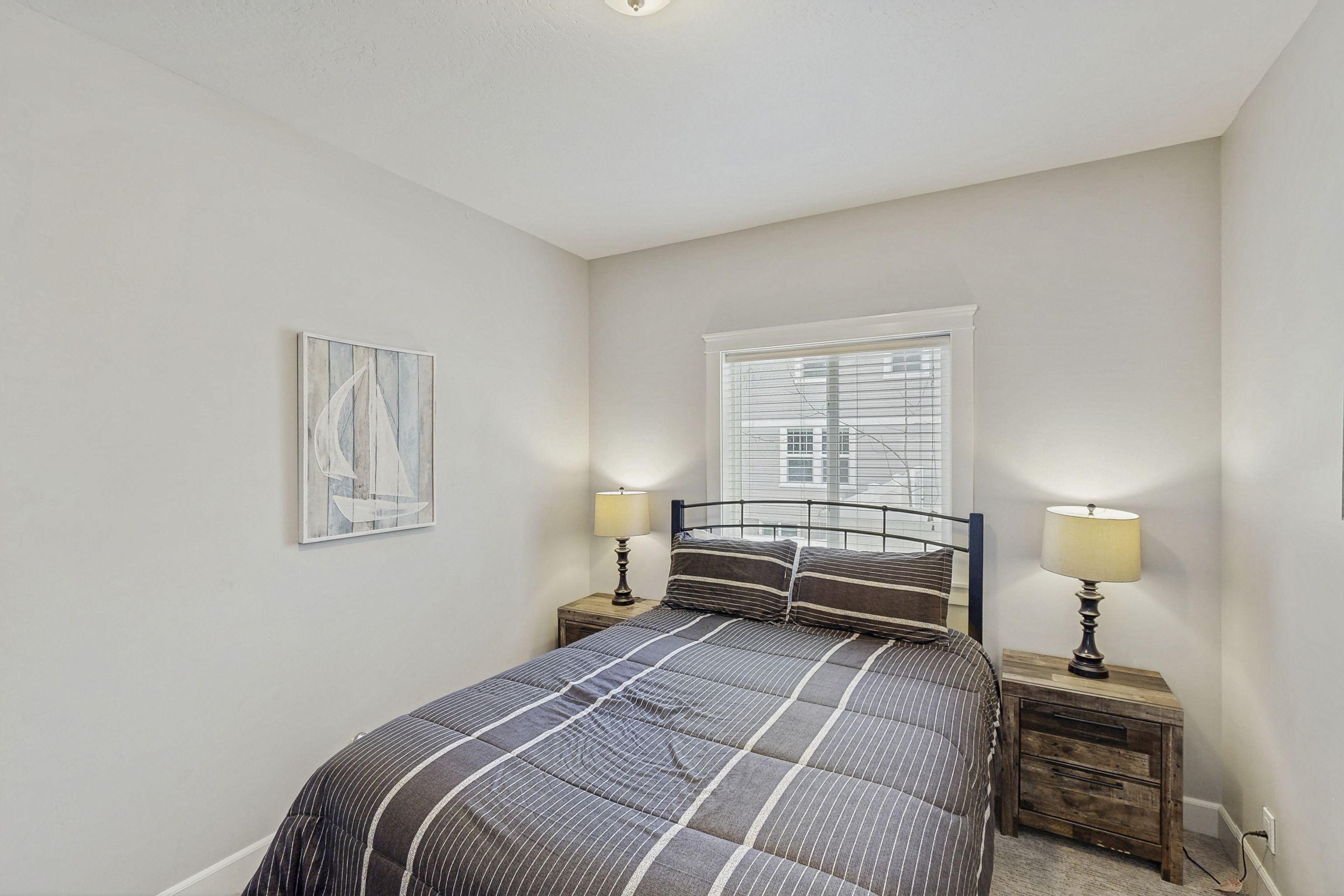 A bedroom with a bed, two bedside tables with lamps, a window with blinds, and a wall-mounted picture.