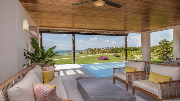 Ein geräumiges Zimmer mit Deckenventilator, einem großen Fenster mit Blick auf einen Golfplatz und den Ozean sowie einer Sitzecke mit Sofa und Sesseln.